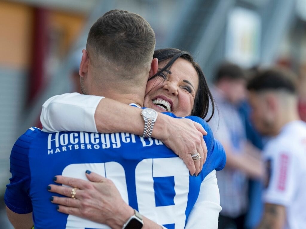 Zwei Personen umarmen sich, eine trägt ein blaues Trikot mit der Aufschrift 'Hochegger Bau HARTBERG' und der Nummer 28