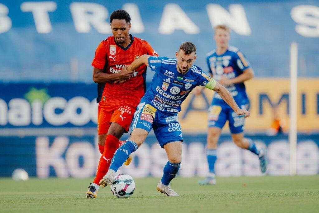 Zwei Fußballspieler in Aktion auf dem Spielfeld, einer in rotem Trikot, der andere in blauem Trikot mit Ball am Fuß, im Hintergrund ein weiterer Spieler in blauem Trikot